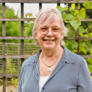 Smiling woman, with white hair. She's wearing glasses and has a fence in the back with green leaves.