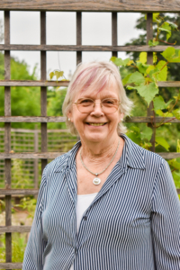 Smiling woman, with white hair. She's wearing glasses and has a fence in the back with green leaves.